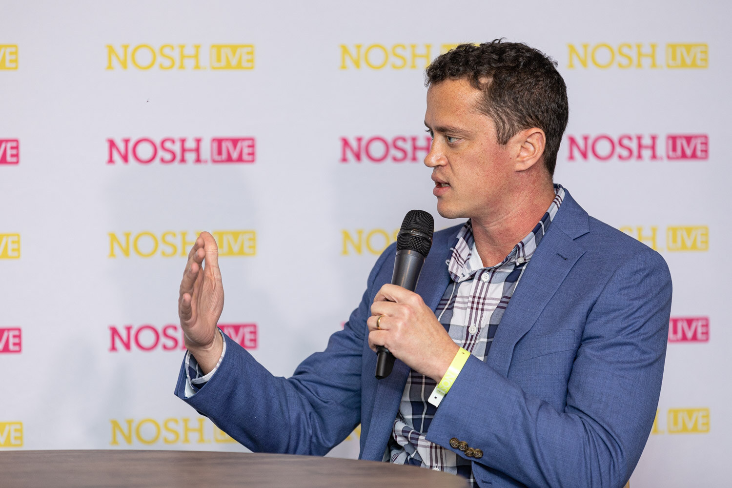 Man in a blue blazer and plaid shirt seen in profile, holding a microphone and raising his other hand, in front of a yellow and pink Nosh Live backdrop.