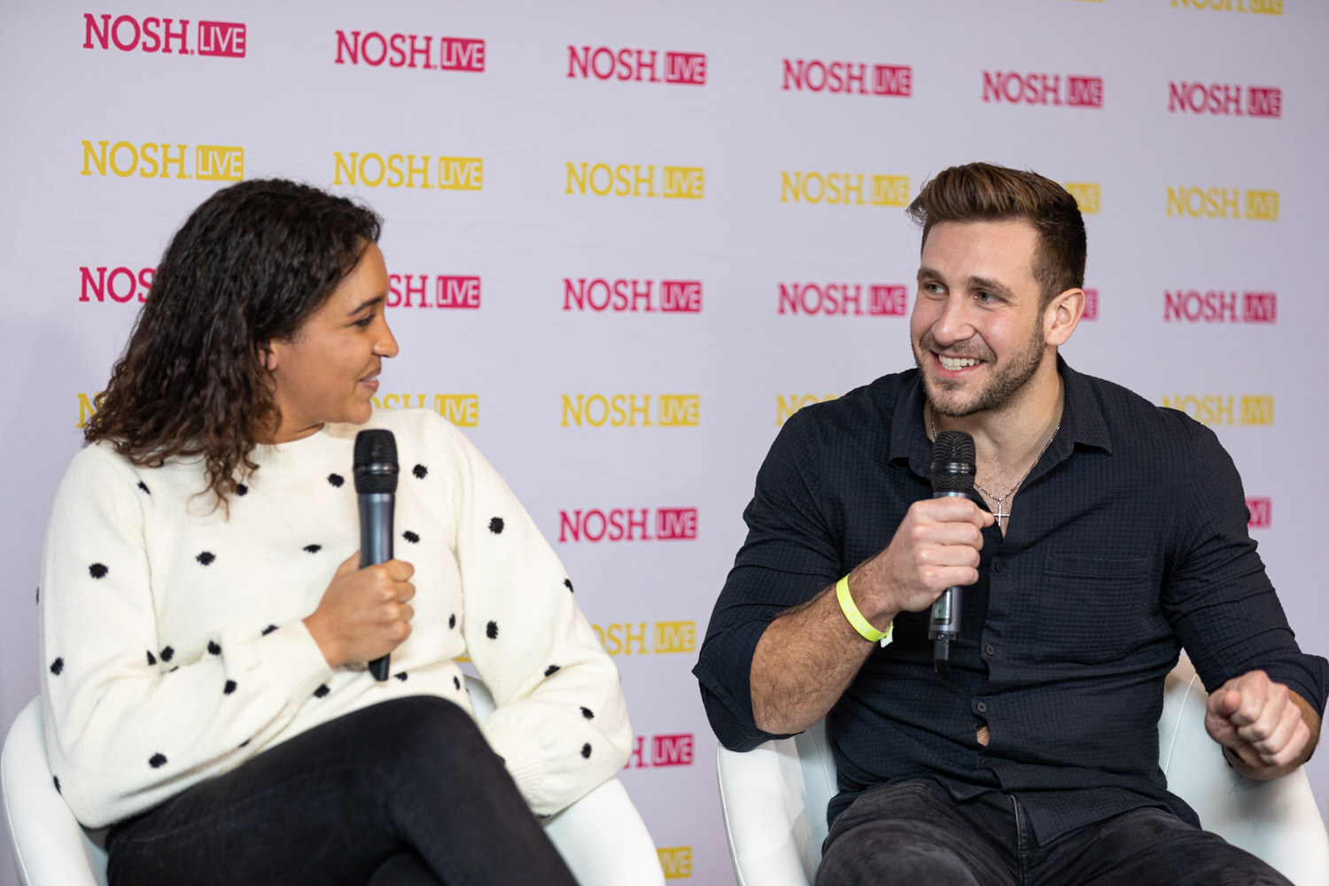 Woman with curly dark hair in a cream polka-dot sweater and a man in a dark button-up shirt, both holding microphones and smiling, seated on white chairs in front of a pink and yellow Nosh Live backdrop.