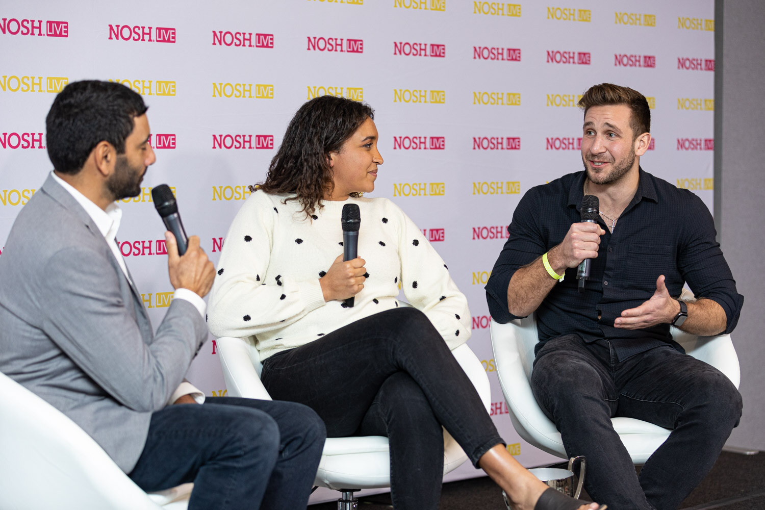 Man in a grey blazer holding a microphone, a woman in a cream polka-dot sweater and a man in a dark shirt seated on white chairs in front of a Nosh Live backdrop.