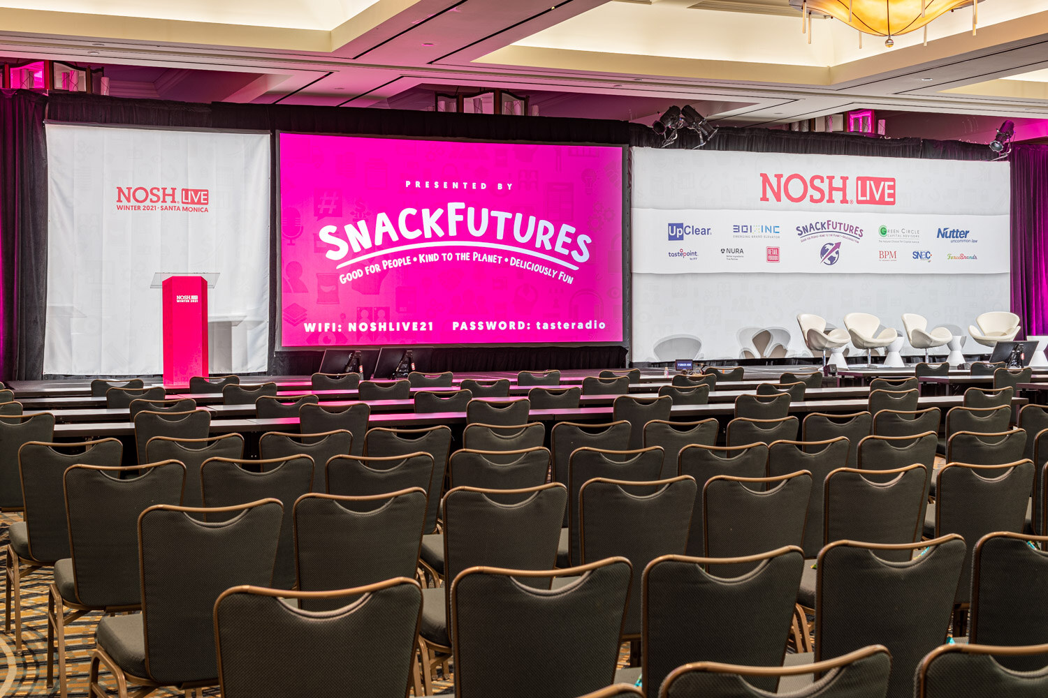 Wide view of a ballroom with rows of dark chairs facing a stage with a pink podium, a pink SnackFutures screen showing Wi-Fi details, a Nosh Live sponsor wall and white chairs.