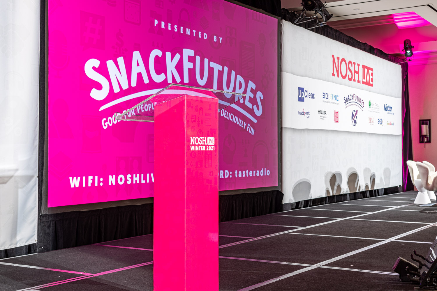 Pink podium labeled Nosh Live Winter 2021 on a stage in front of a large pink screen reading Presented by SnackFutures, with the sponsor wall and white chairs at the right.
