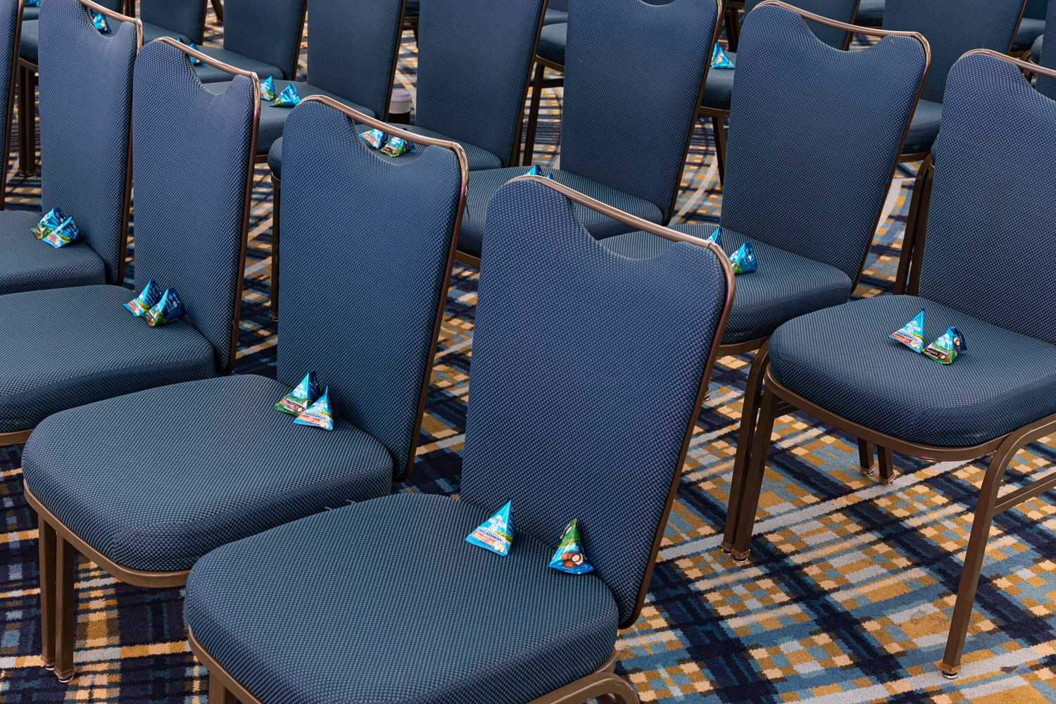 Rows of blue upholstered chairs on a patterned carpet, each with a few small foil-wrapped pyramid-shaped candies on the seat.
