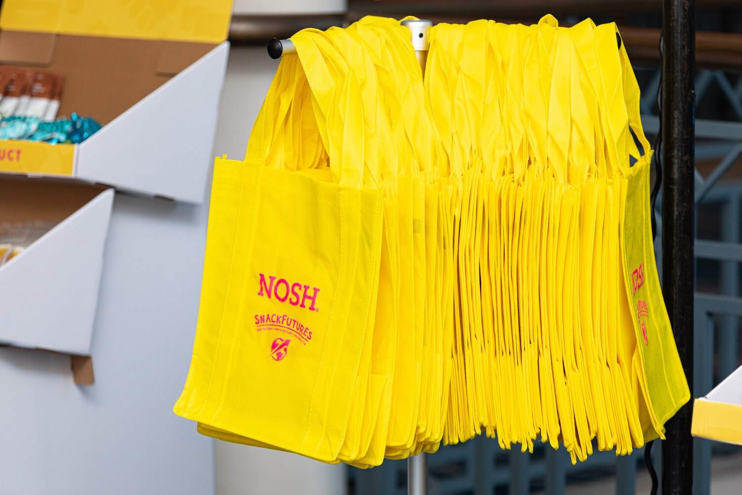 Dozens of yellow reusable tote bags printed with Nosh and SnackFutures hanging together on a metal stand, next to a cardboard display of snacks.