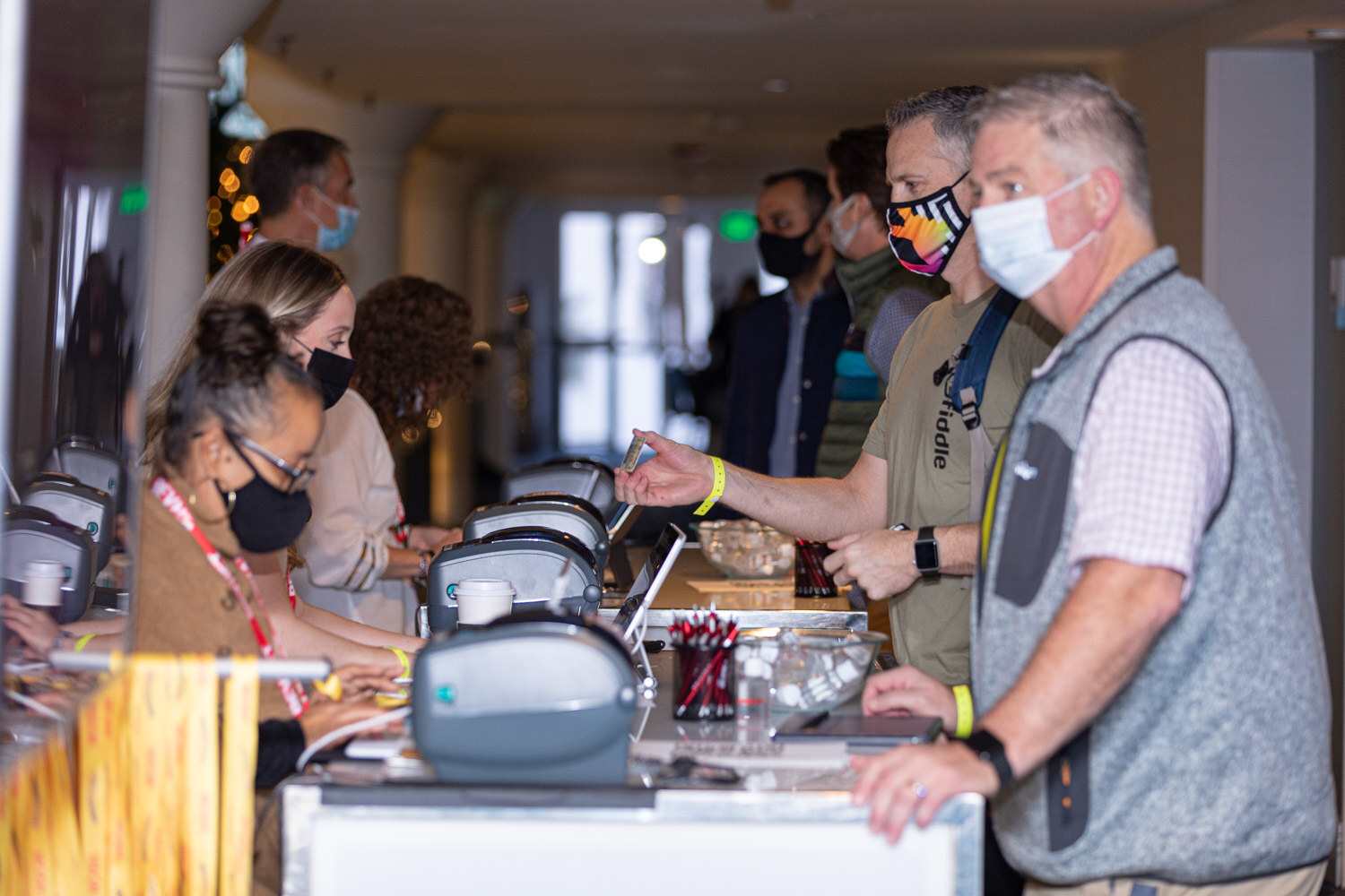 Row of badge printers on a long counter with staff in masks typing at laptops and two attendees in masks, one in a grey vest, waiting in line.