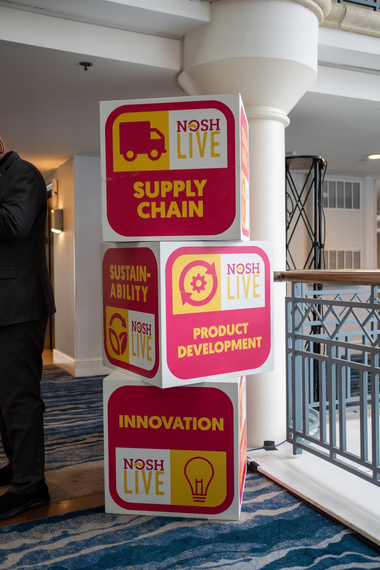 Three stacked pink and yellow cubes labeled Supply Chain, Sustainability, Product Development and Innovation beside a white column and a balcony railing.