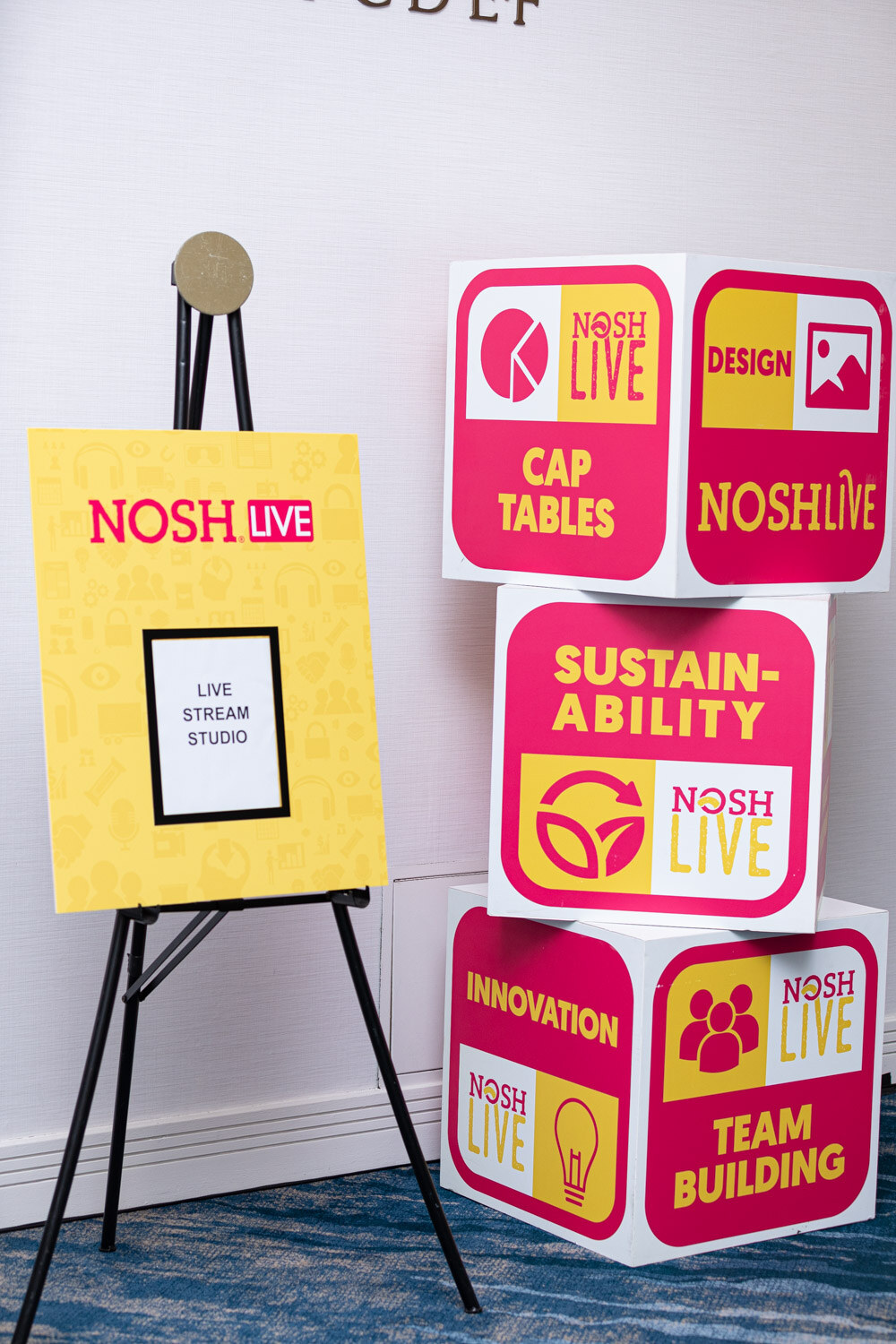 Yellow easel sign reading Nosh Live Live Stream Studio beside a stack of pink and yellow cubes labeled Cap Tables, Design, Sustainability, Innovation and Team Building.