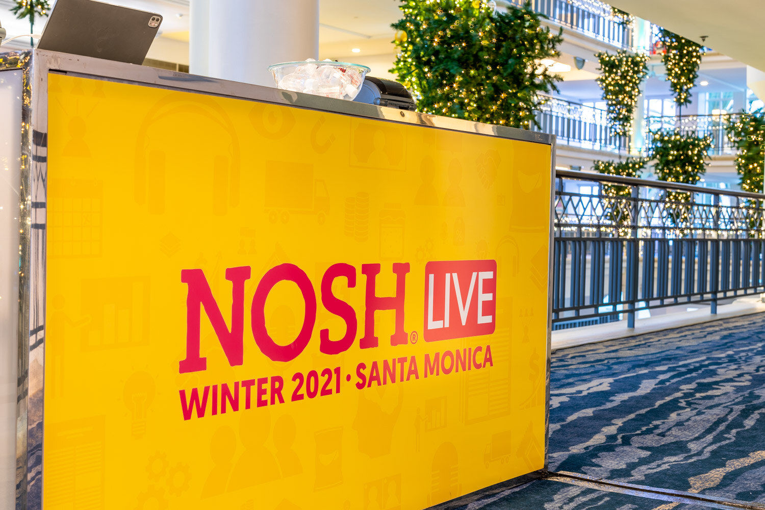 Large yellow panel printed with Nosh Live Winter 2021 Santa Monica on a table with a laptop and a bowl of ice, with hanging lit garlands and a balcony railing behind.