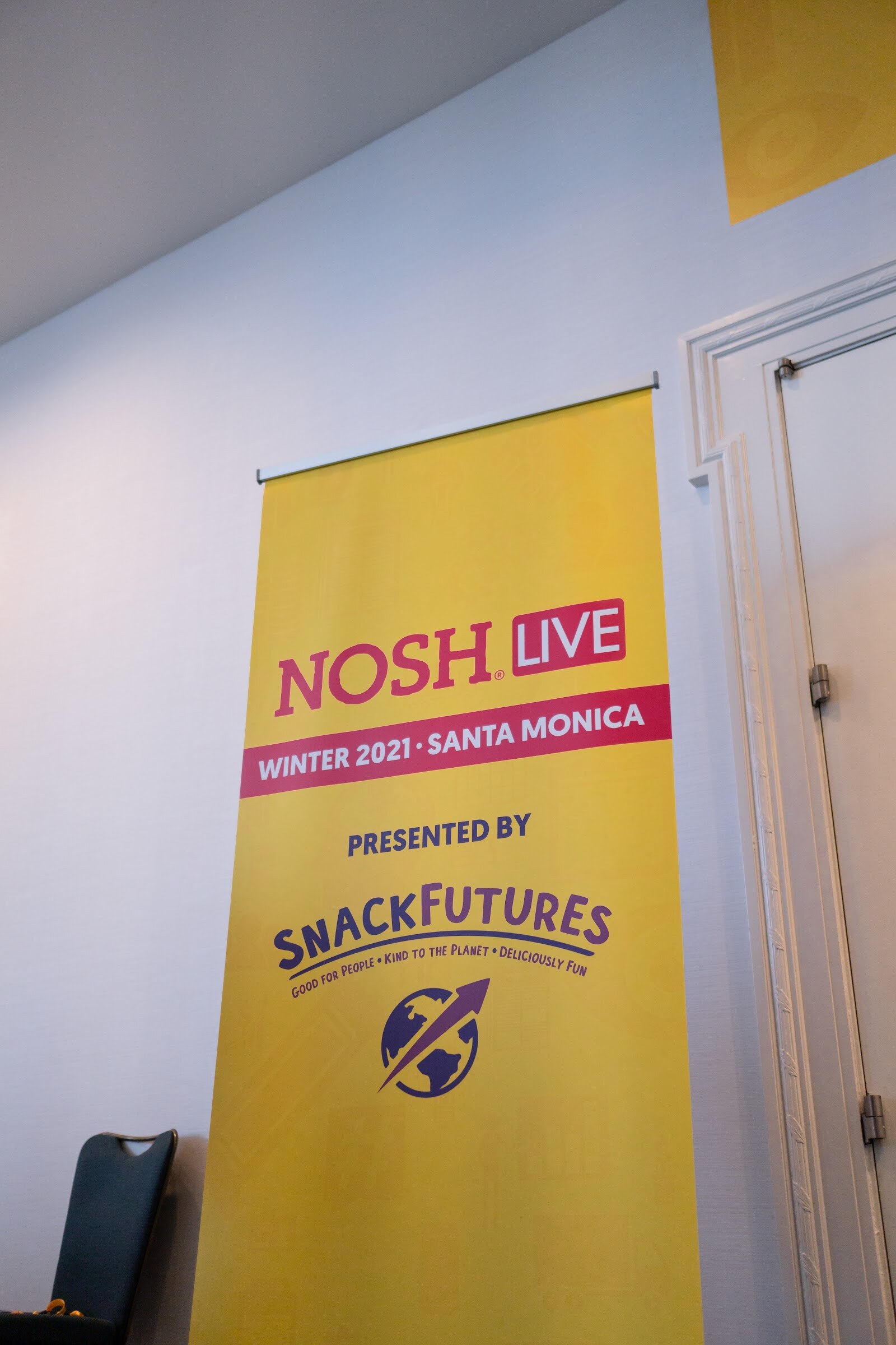 Tall yellow banner with the Nosh Live logo, Winter 2021 Santa Monica and Presented by SnackFutures hanging on a white wall beside a door.