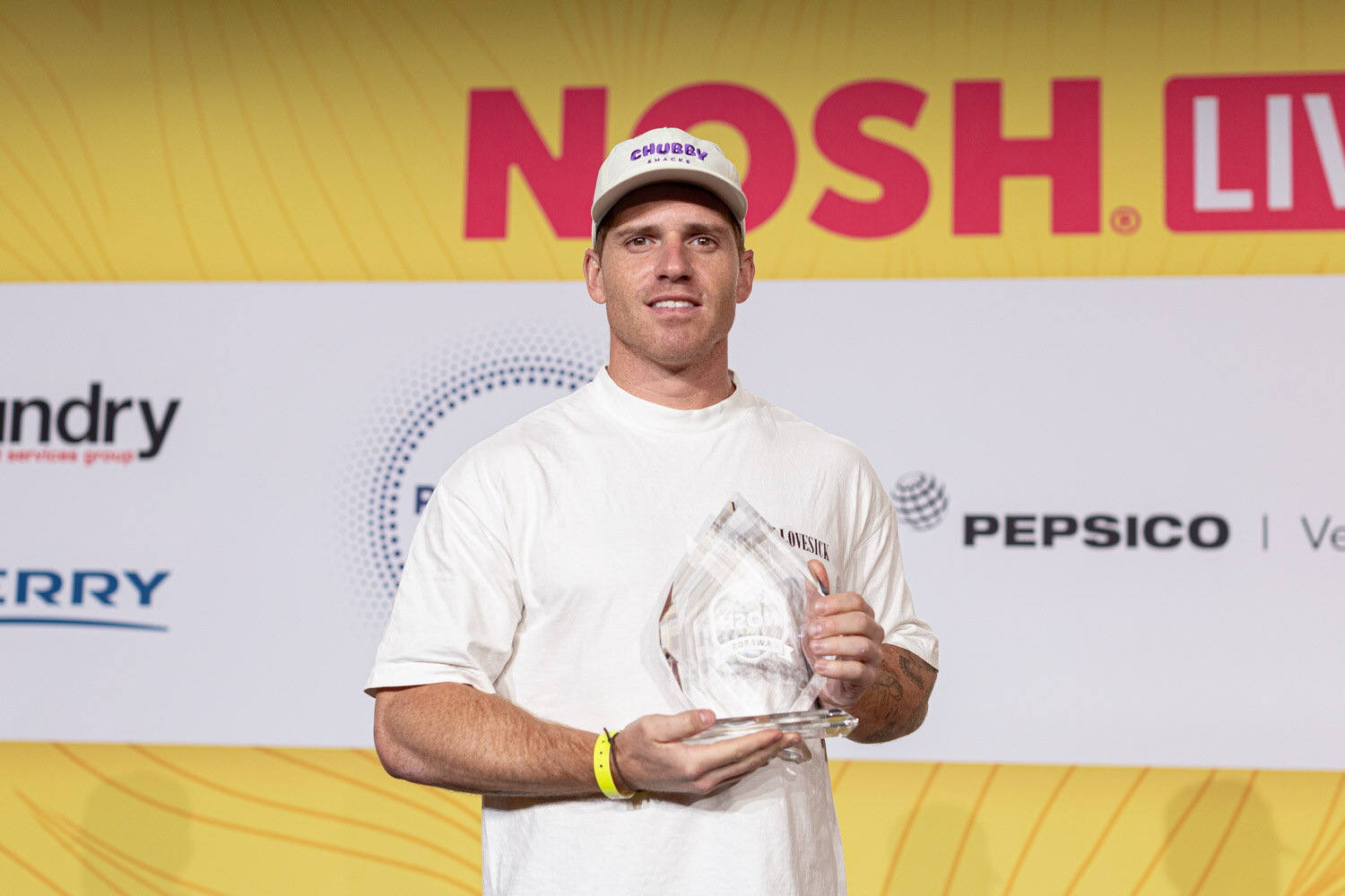 Man in a white t-shirt and cap holding a clear glass trophy in front of the Nosh Live backdrop.