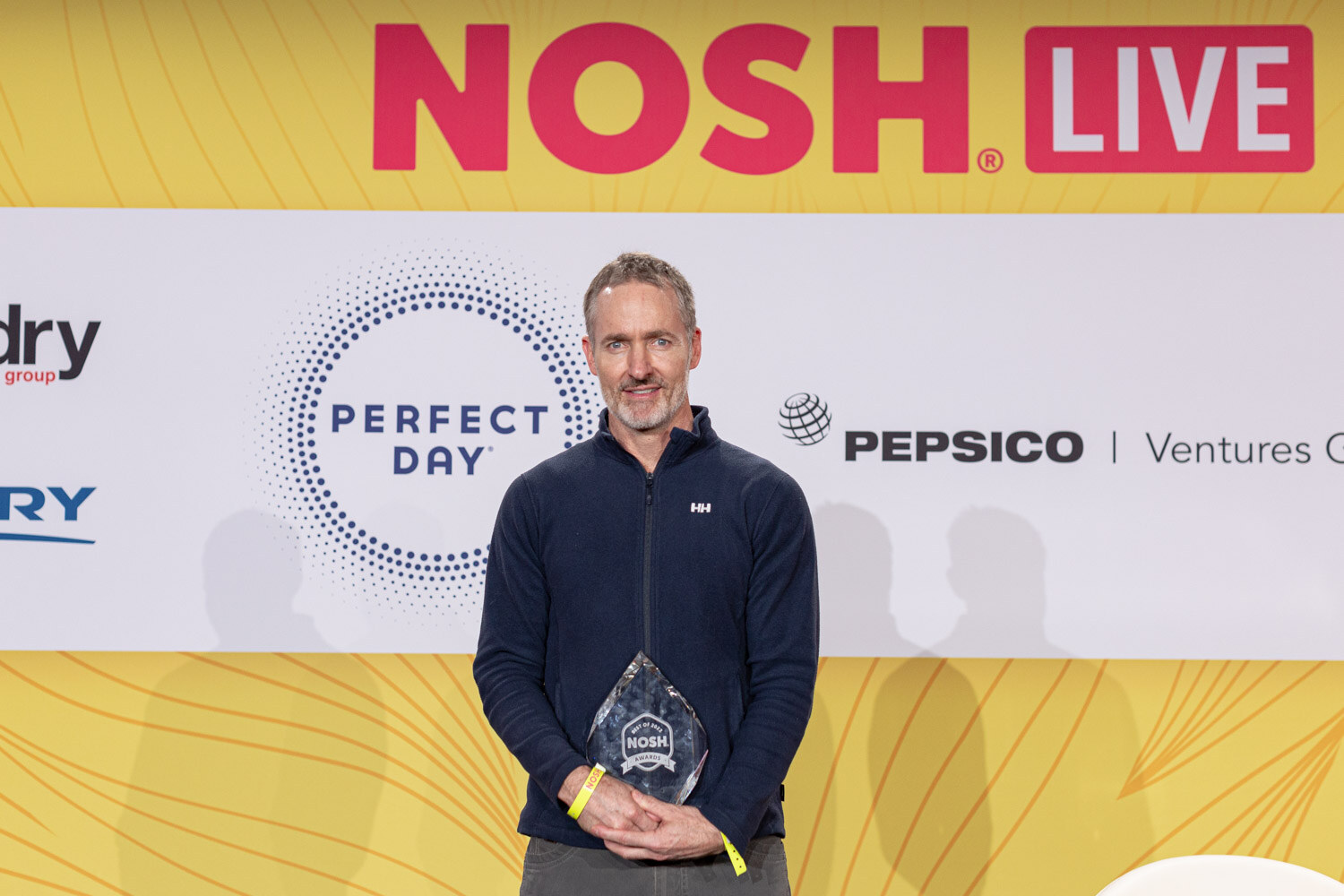 Man in a navy fleece holding a small glass trophy in front of a Perfect Day and Pepsico Ventures Group backdrop.