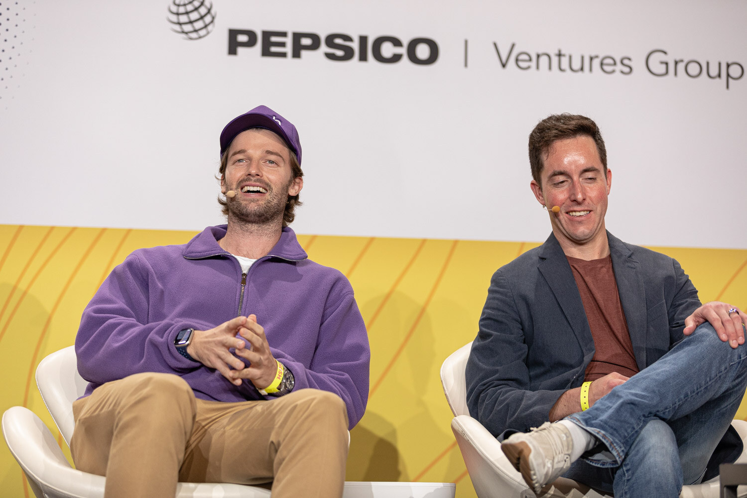 Man in a purple pullover and cap and a man in a navy blazer seated on stage, laughing.