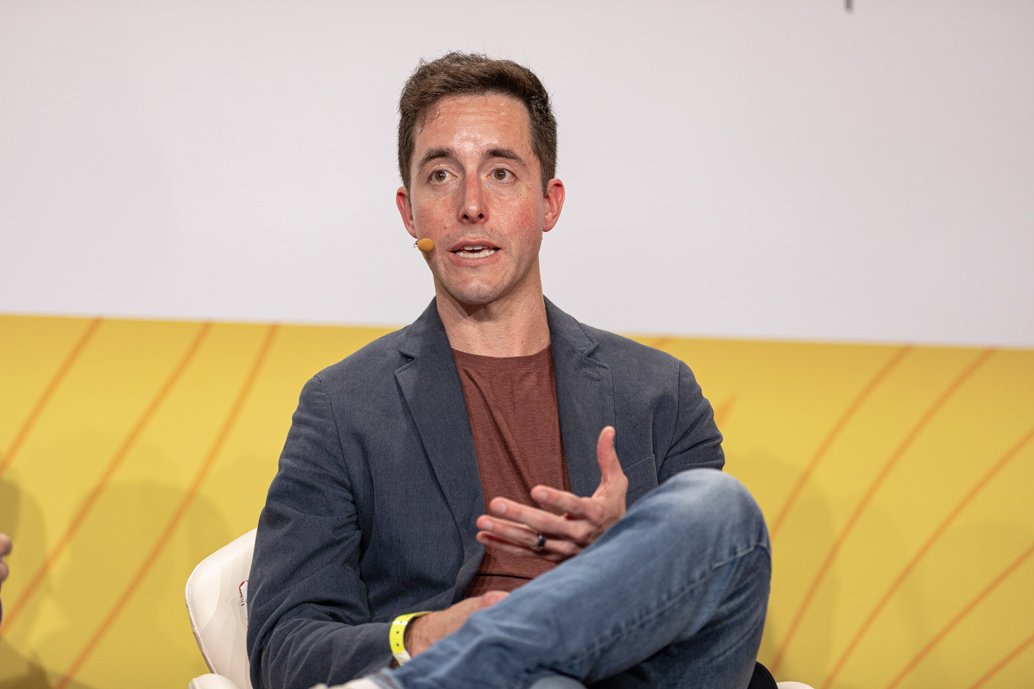 Man in a dark blazer and rust t-shirt gesturing while seated on stage.