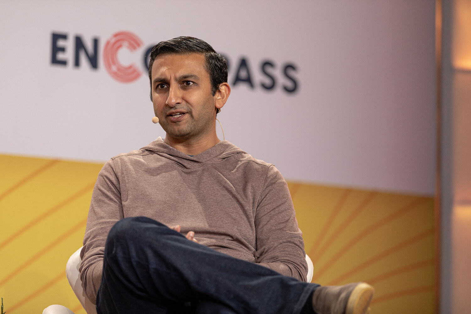 Man in a gray-brown hoodie seated with crossed legs in front of an Encompass logo.