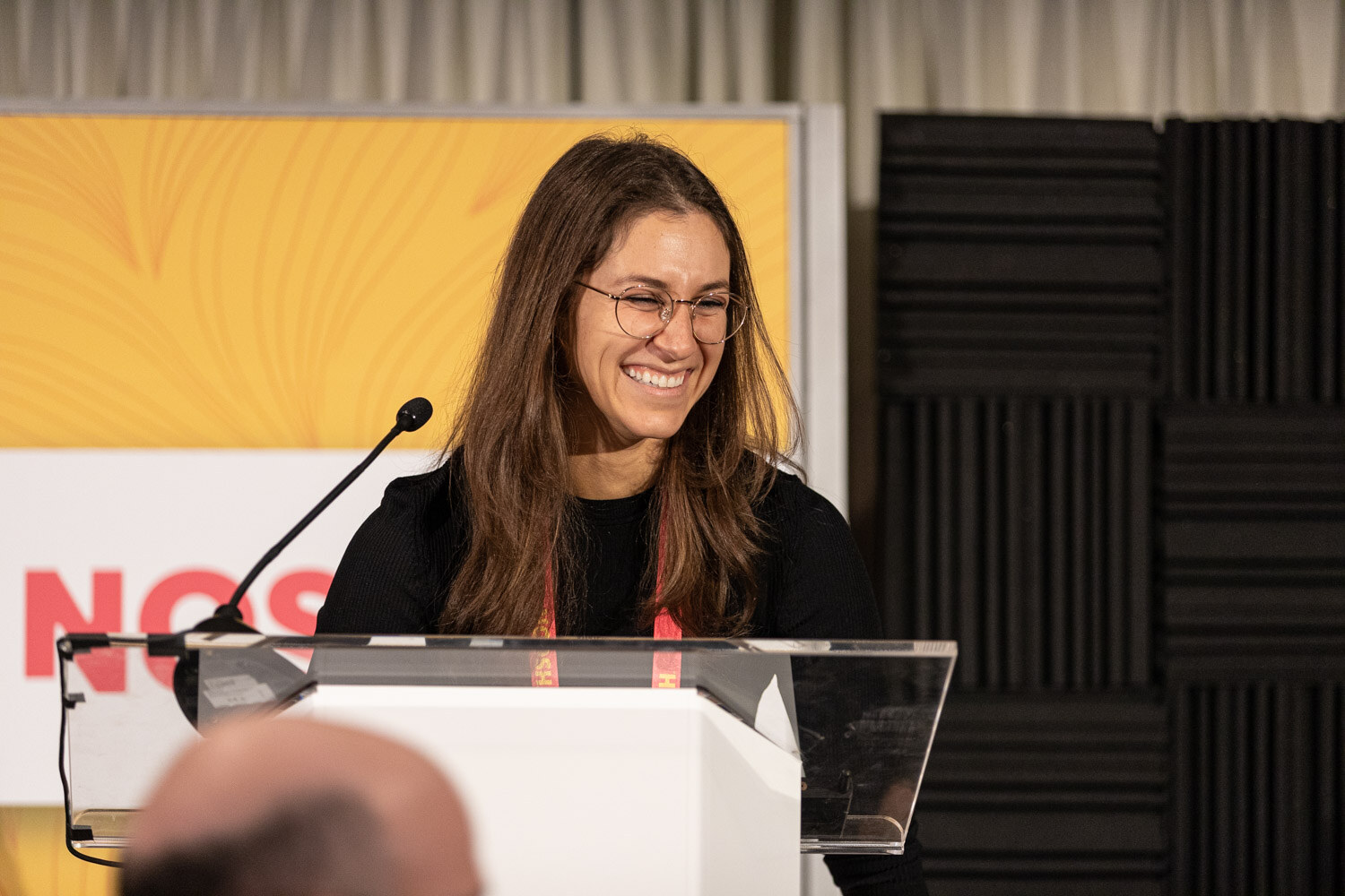 Smiling woman with glasses behind a clear lectern with a Nosh Live banner behind her.