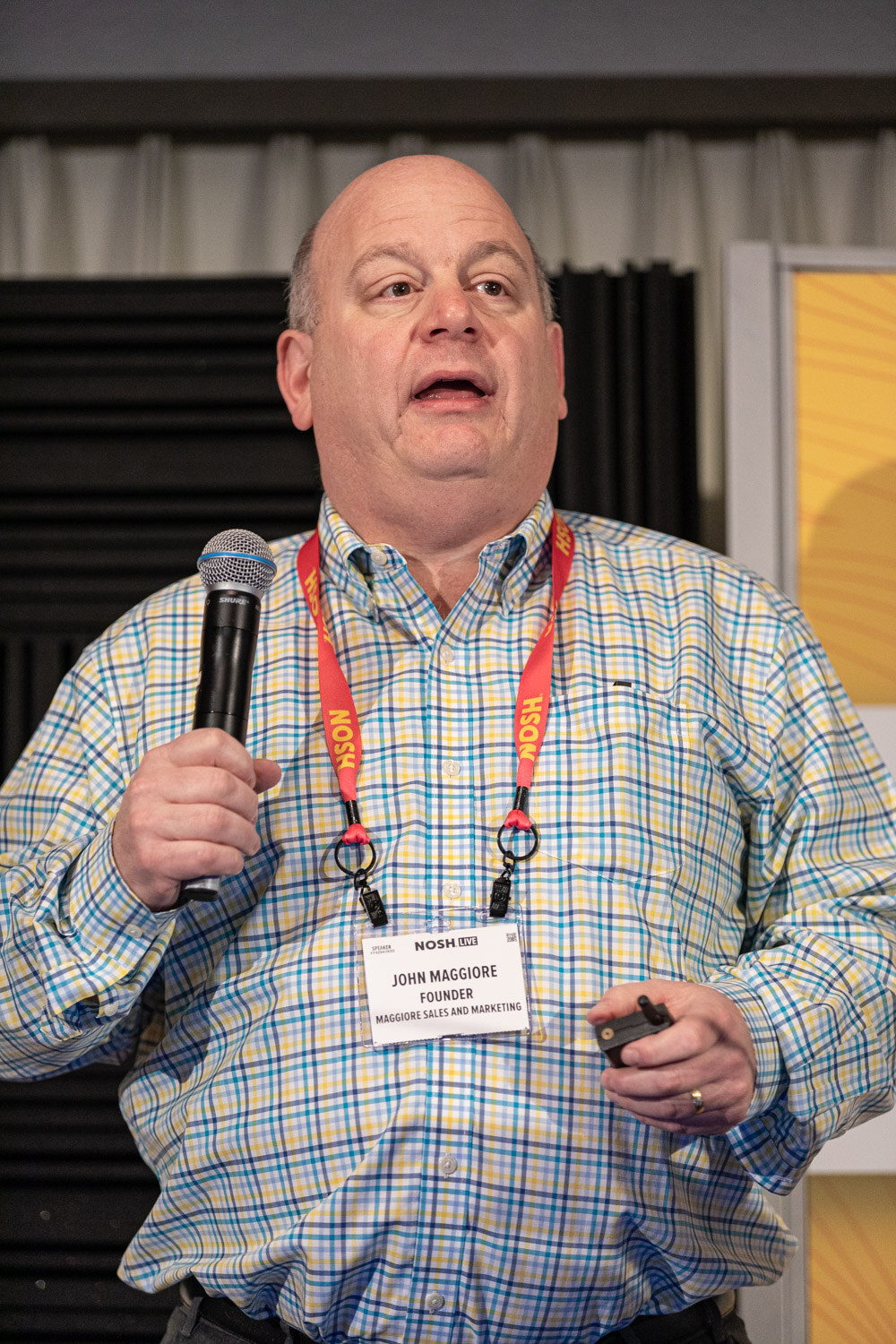 Man in a plaid shirt with a lanyard holding a microphone and presentation clicker.