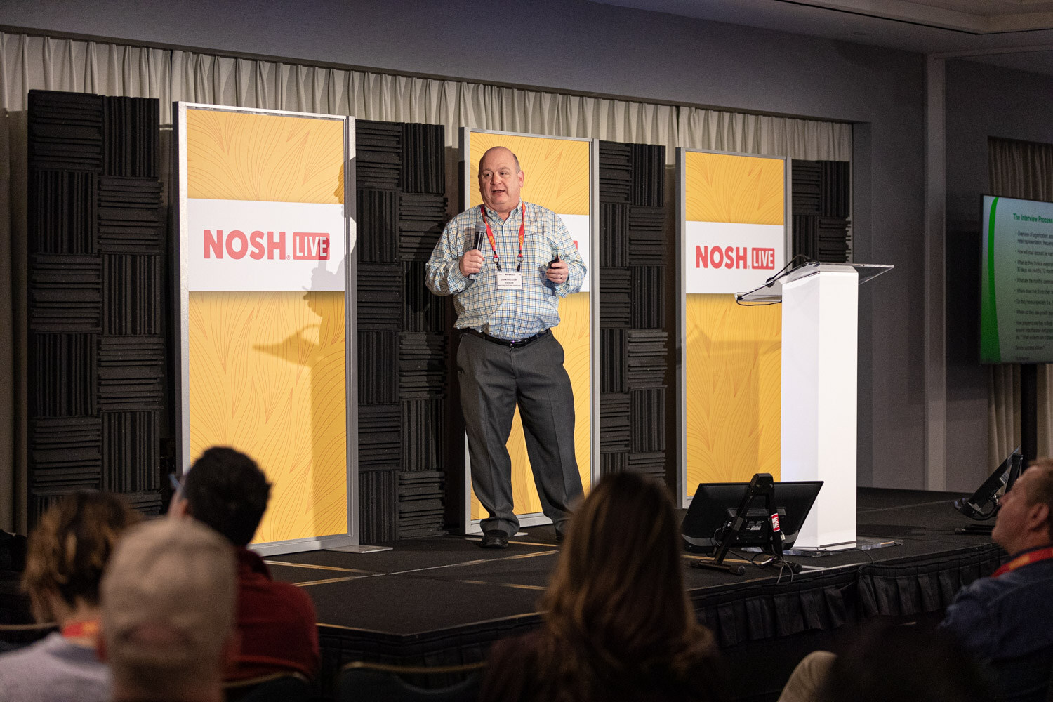 Man in a plaid shirt presenting on a low stage with yellow Nosh Live panels, audience heads in the foreground.