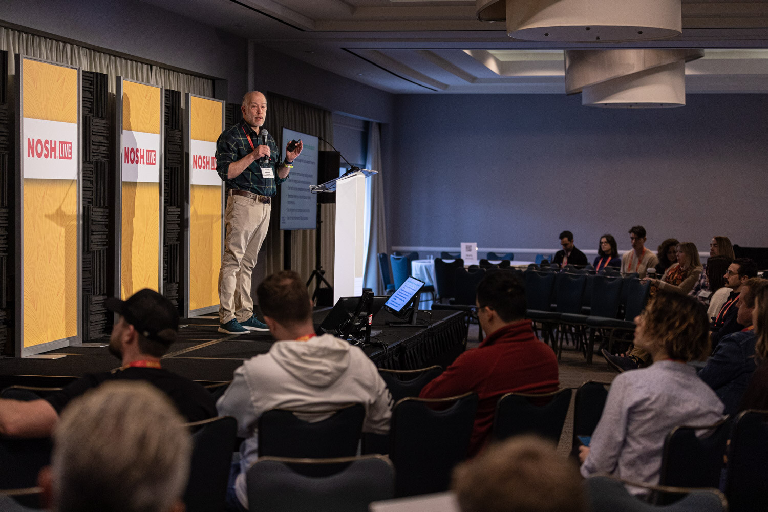 Man standing on a stage beside a lectern while seated attendees watch.