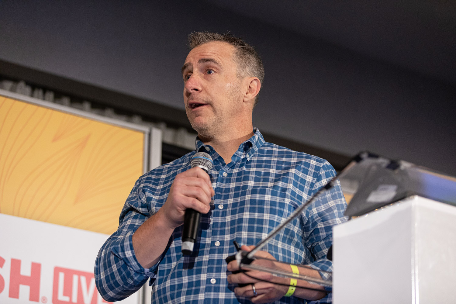 Man in a blue plaid shirt holding a microphone near a lectern with a partial Nosh Live sign.
