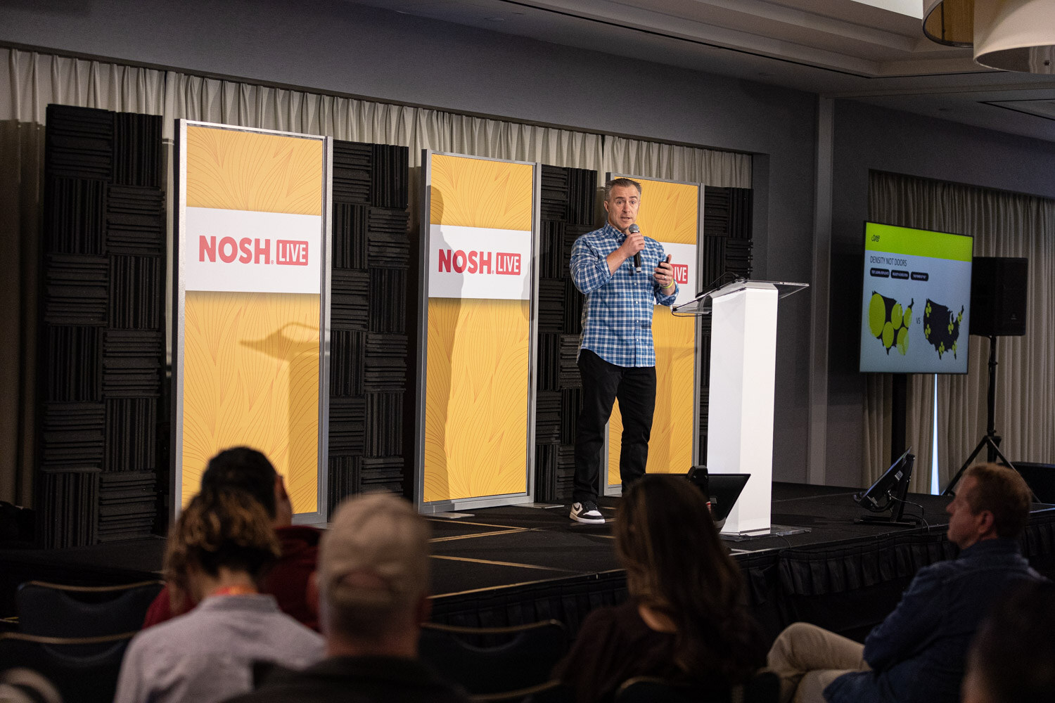 Man in a plaid shirt on a small stage with a screen showing two maps and Nosh Live banners.