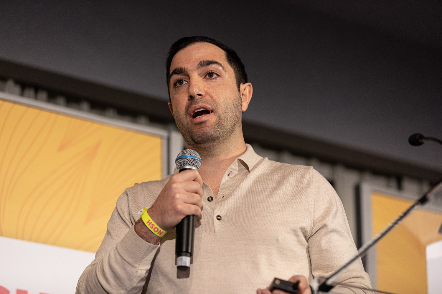 Man in a cream henley shirt with a yellow wristband speaking into a microphone.