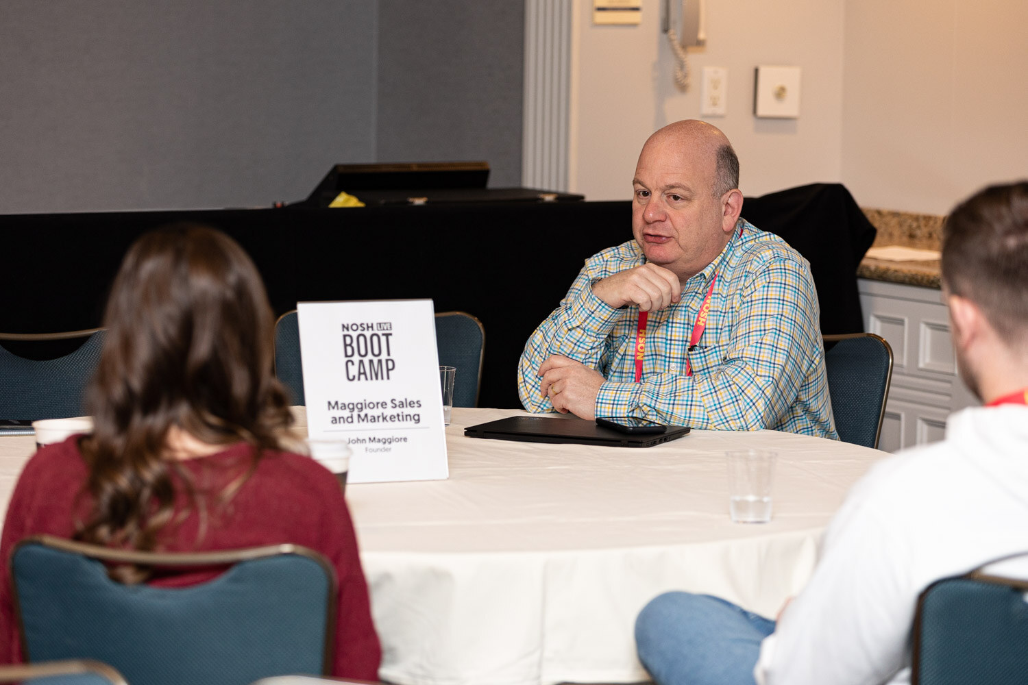 Man in a light plaid shirt seated at a round table beside a table sign reading Boot Camp, Maggiore Sales and Marketing.