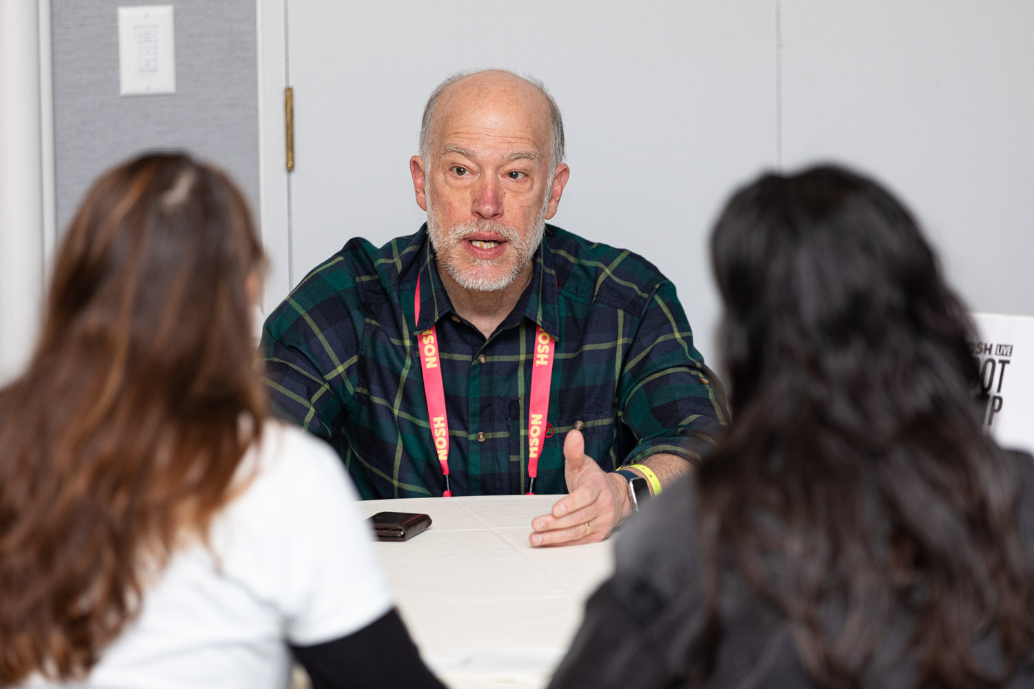Gray-haired man in a green plaid shirt gesturing while speaking to two women seated with their backs to the camera.