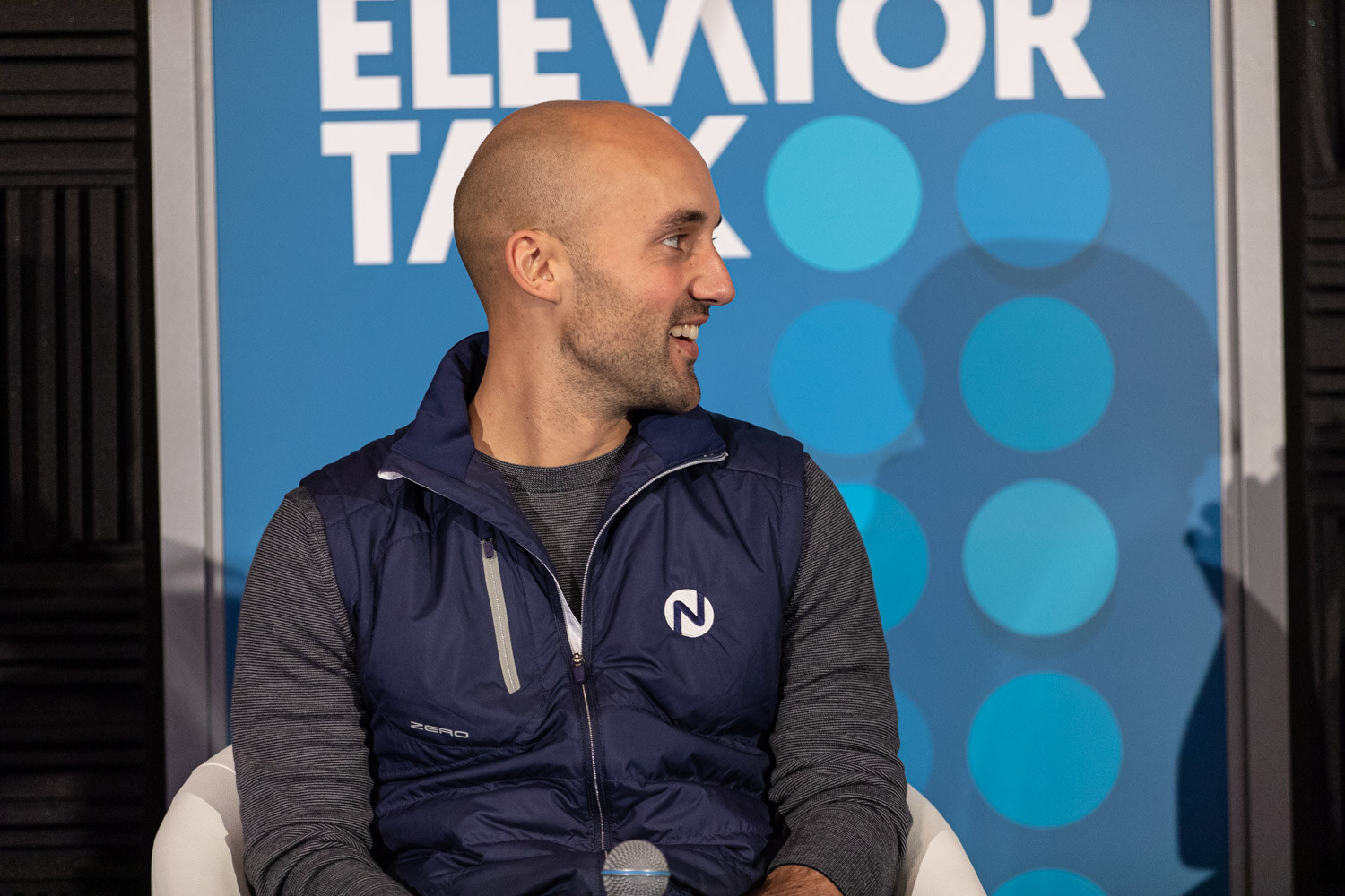 Bald man in a navy vest seated in front of an Elevator Talk banner, smiling toward the side.