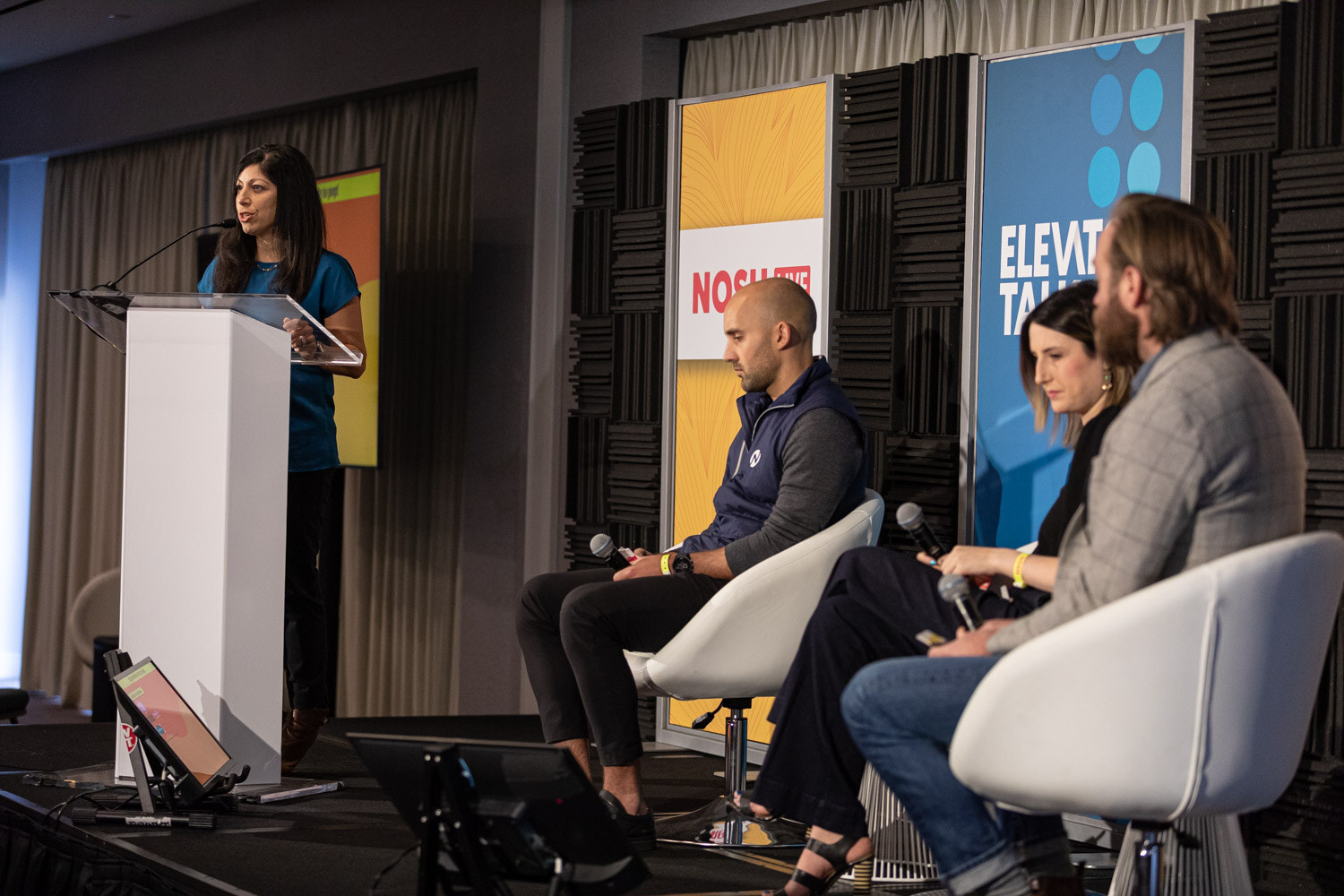 Woman at a white lectern with three seated panelists and Elevator Talk and Nosh Live banners behind them.