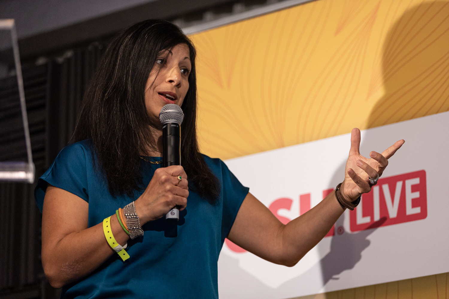 Woman in a teal top holding a microphone and pointing with her other hand in front of a Nosh Live banner.
