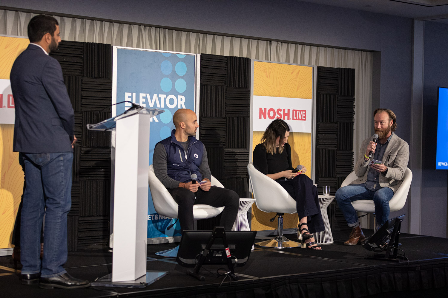 Man at a white lectern facing three seated panelists in front of Elevator Talk and Nosh Live banners.
