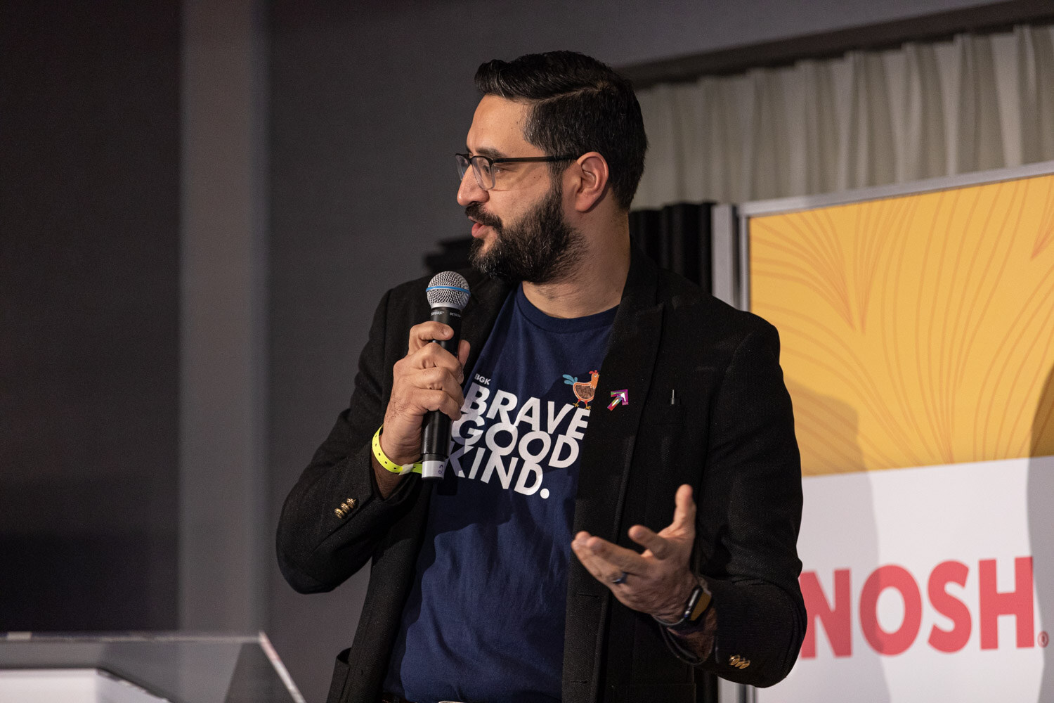 Bearded man with glasses holding a microphone, wearing a navy shirt that reads Brave Good Kind.