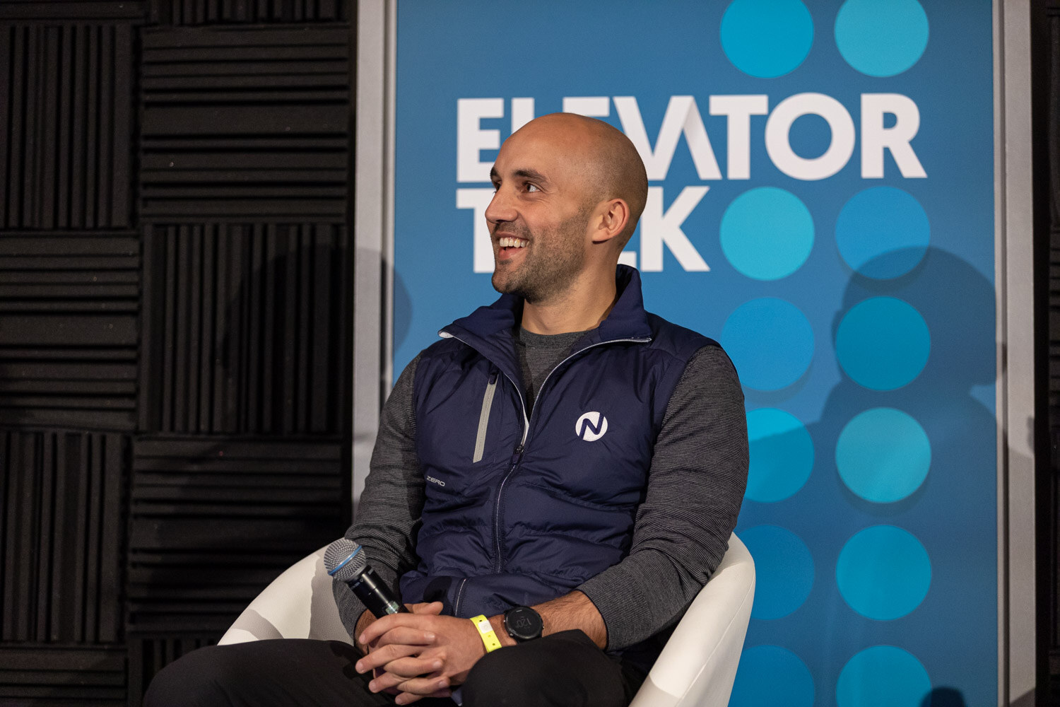 Bald man in a navy vest laughing, seated in front of an Elevator Talk banner.