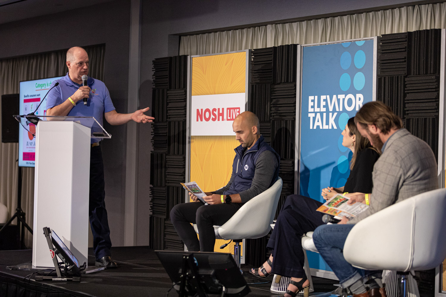 Man in a blue polo at a lectern beside a screen, with three panelists seated nearby.
