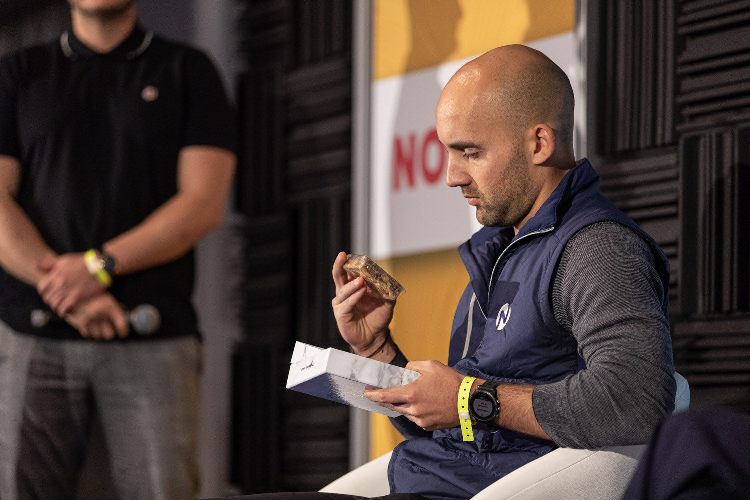 Man in a navy vest looking at a snack bar and its package while another person stands beside him.
