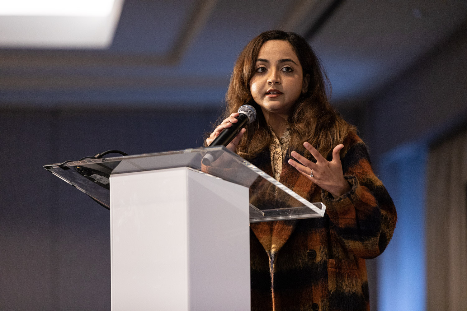 Woman in a brown plaid coat speaking into a microphone at a clear lectern.