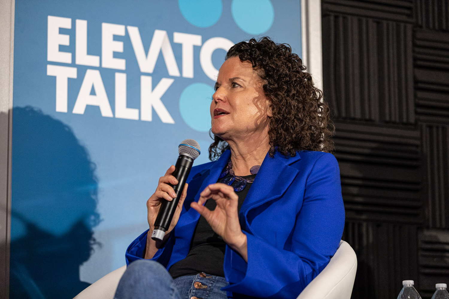 Woman with curly dark hair in a blue blazer holding a microphone, Elevator Talk banner behind her.