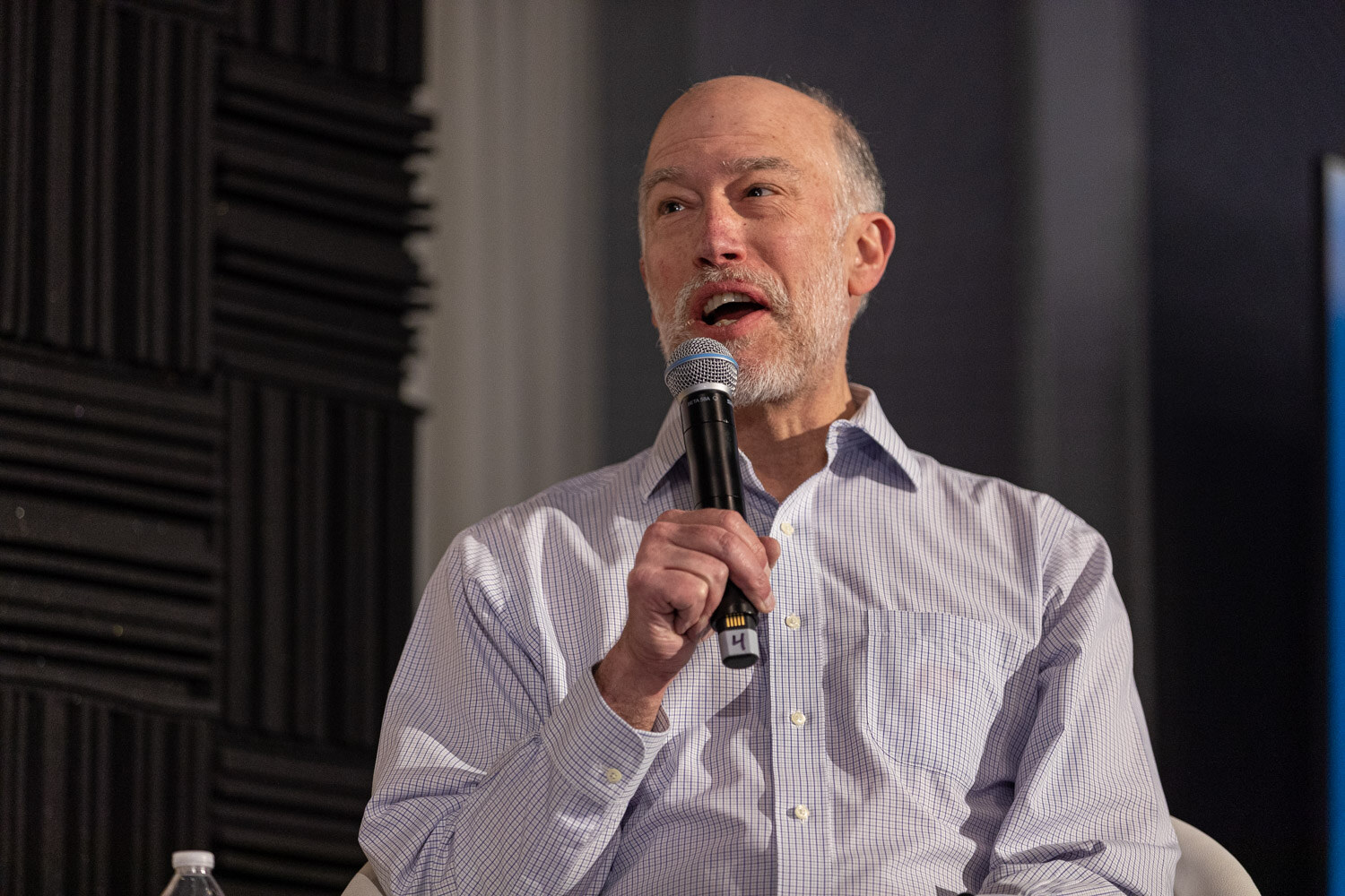 Gray-bearded man in a light checked shirt speaking into a microphone.