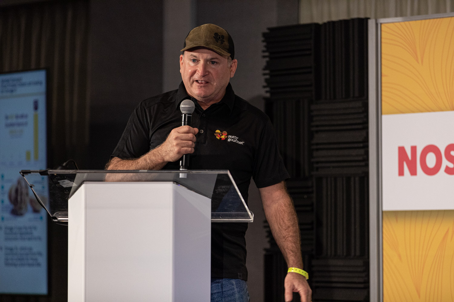 Man in an olive cap and black polo with a logo, holding a microphone behind a lectern.