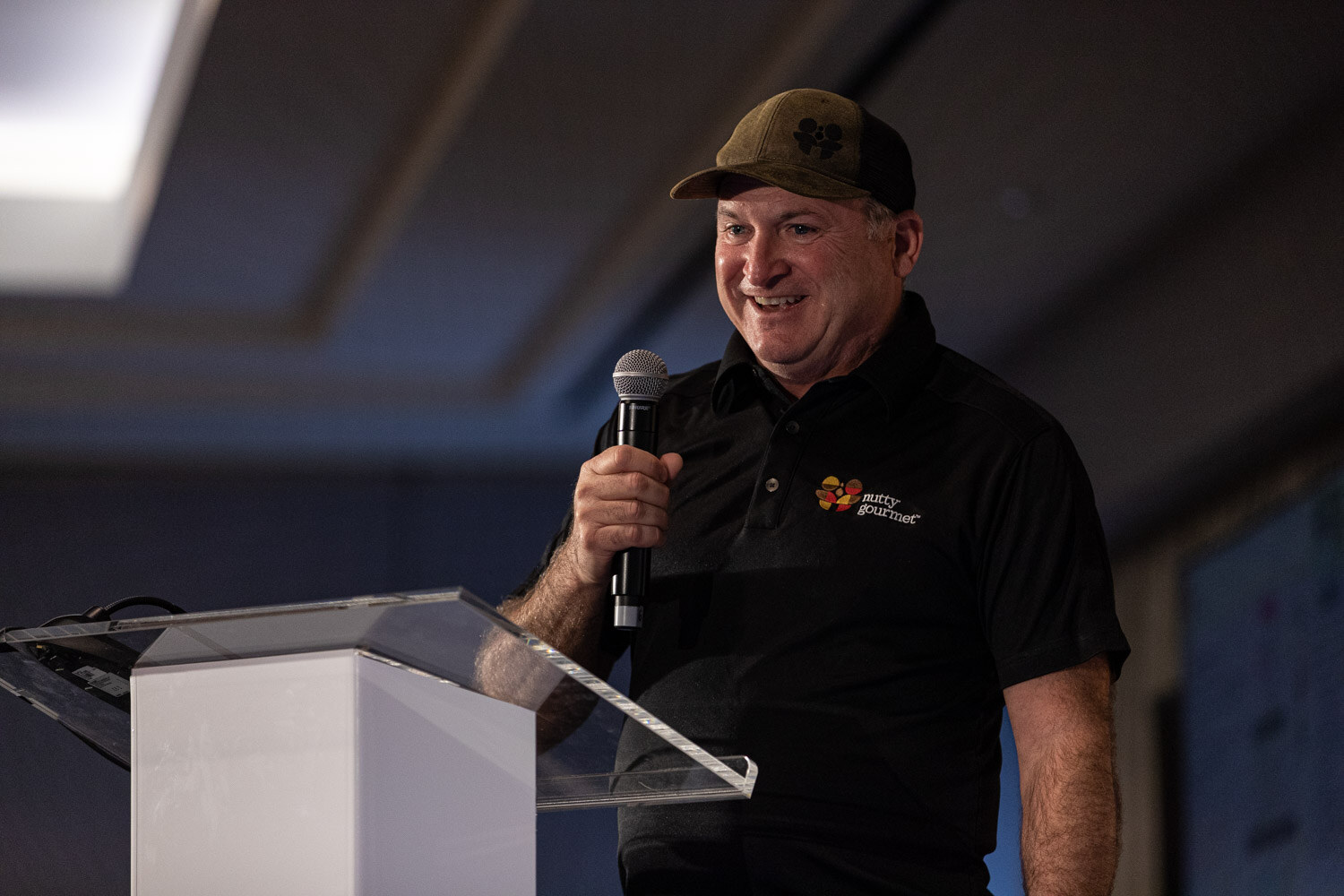 Smiling man in a cap and black polo holding a microphone near a lectern.