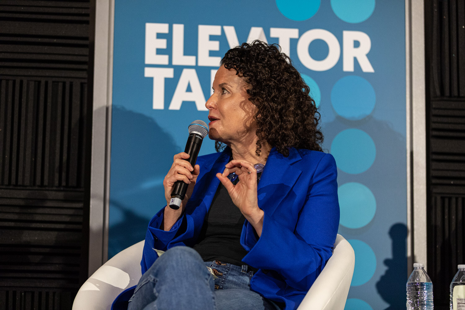 Woman in a blue blazer seated and speaking into a microphone in front of an Elevator Talk banner.