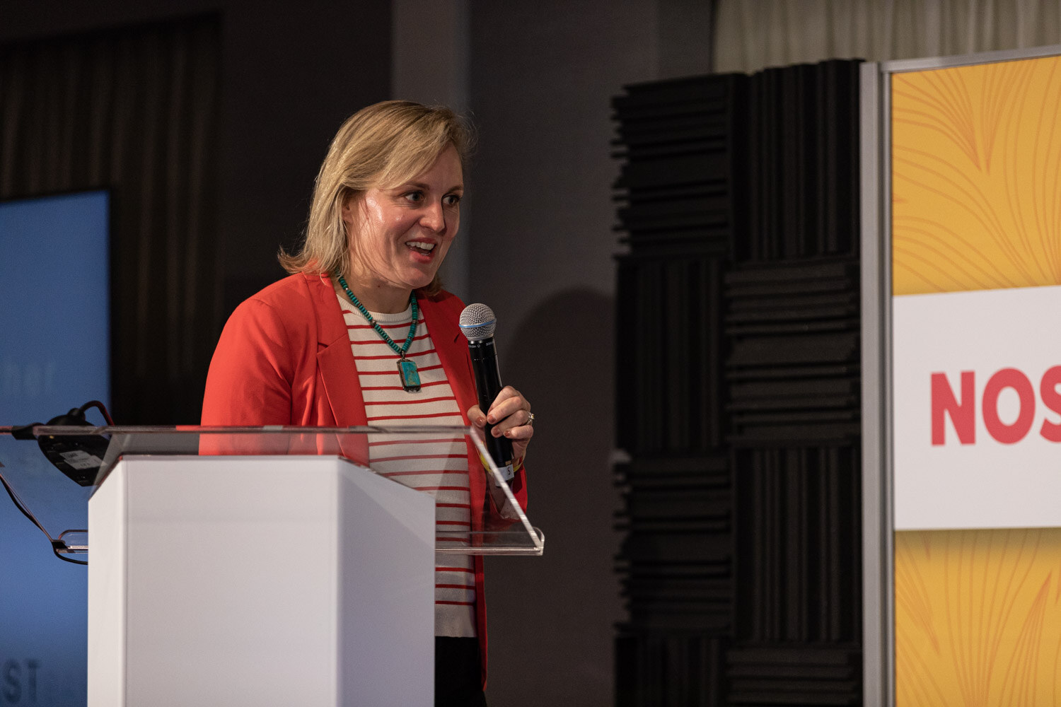 Blond woman in a red blazer and striped shirt holding a microphone at a lectern.