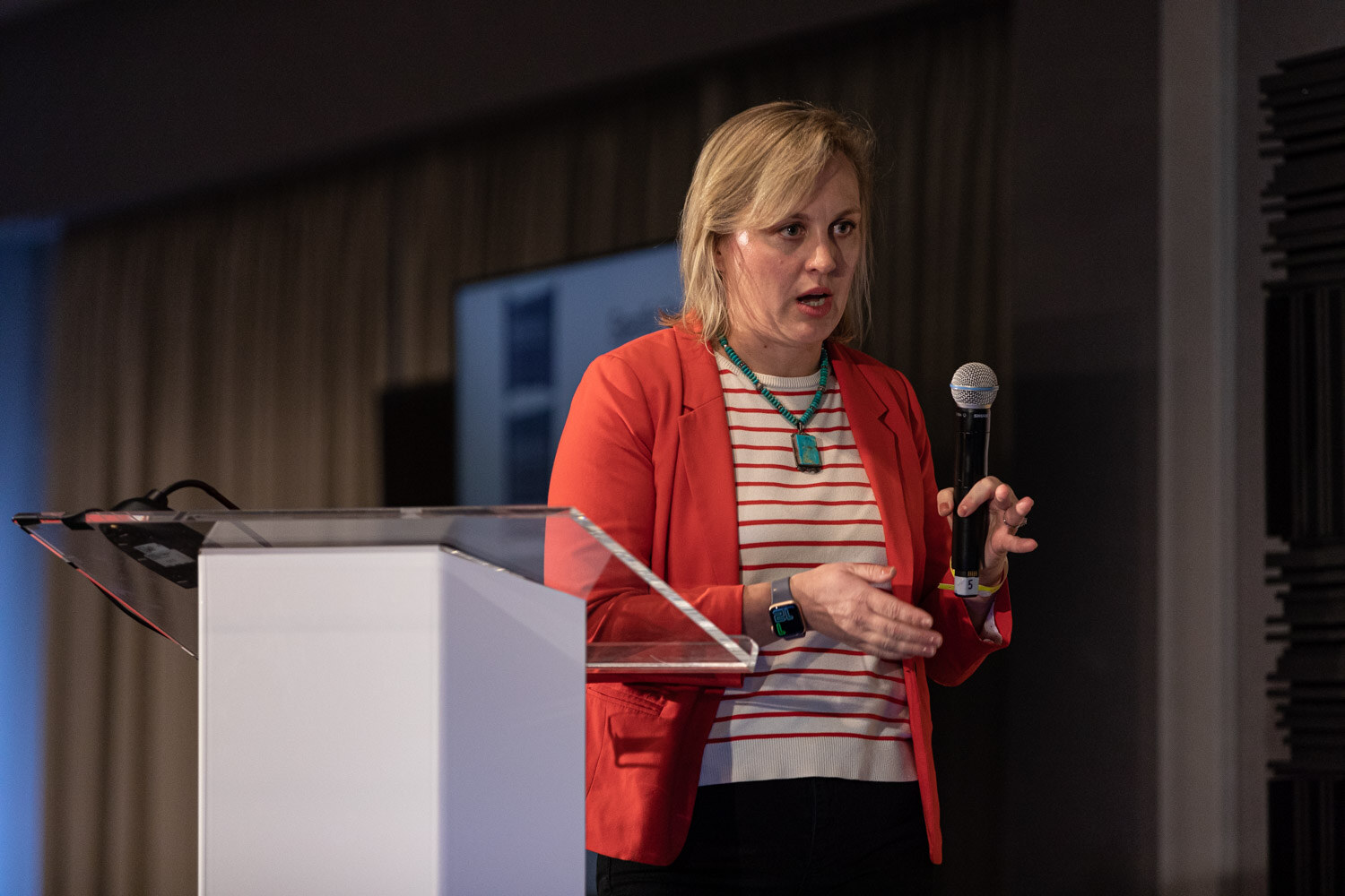 Woman in a red blazer holding a microphone and gesturing with her other hand.