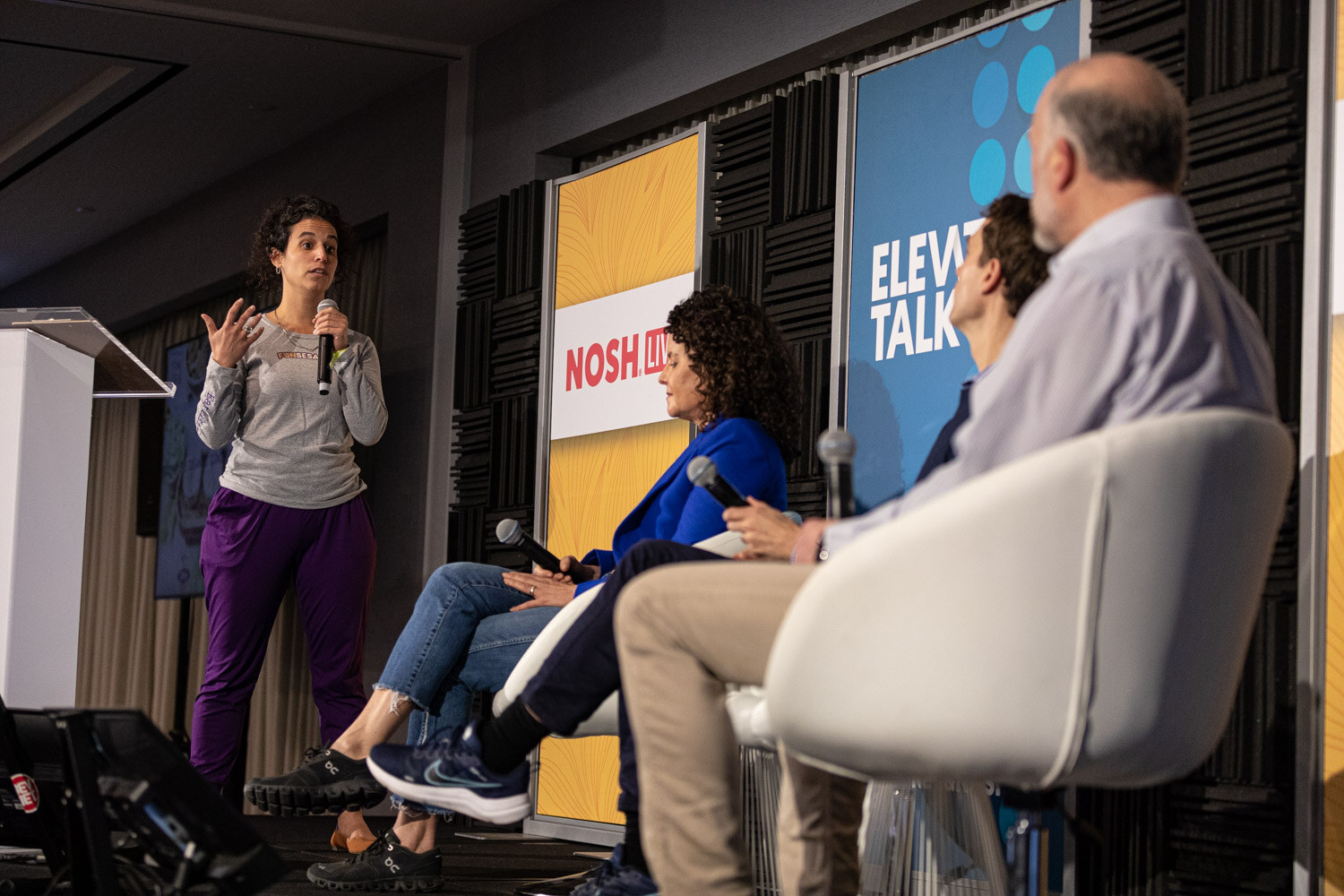 A standing woman gesturing beside three seated panelists in front of Elevator Talk banners.
