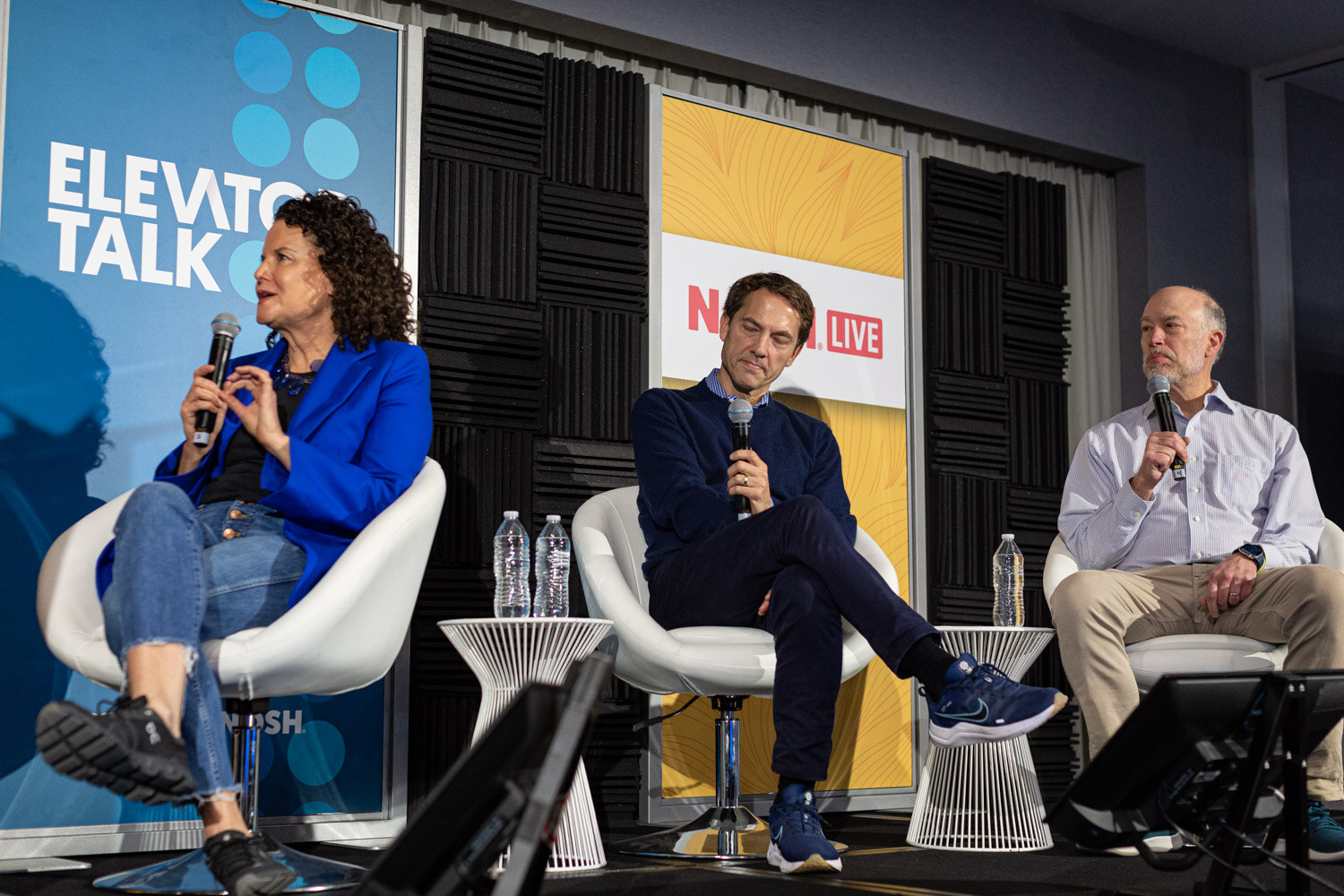 Three seated people with microphones, a woman in a blue blazer and two men, in front of Elevator Talk and Nosh Live banners.