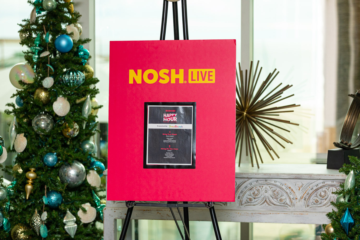 Pink Nosh Live easel sign with a menu card beside a holiday tree with blue and silver ornaments.