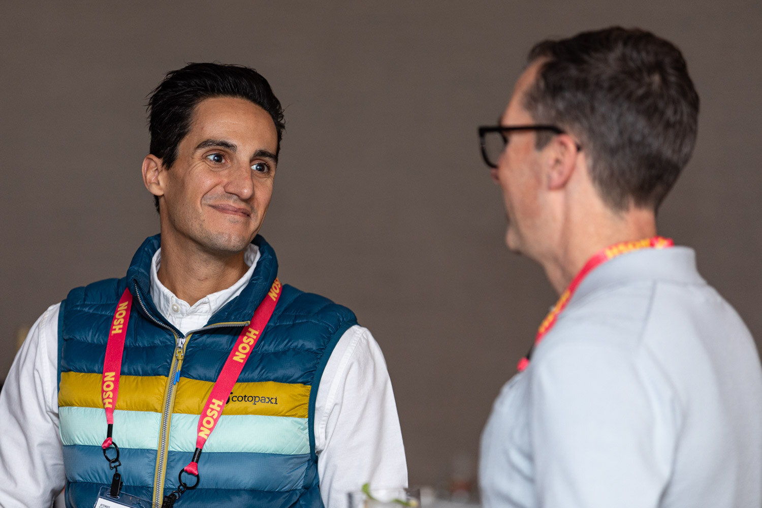 Smiling man in a striped puffer vest talking with another man in profile, both wearing red Nosh lanyards.