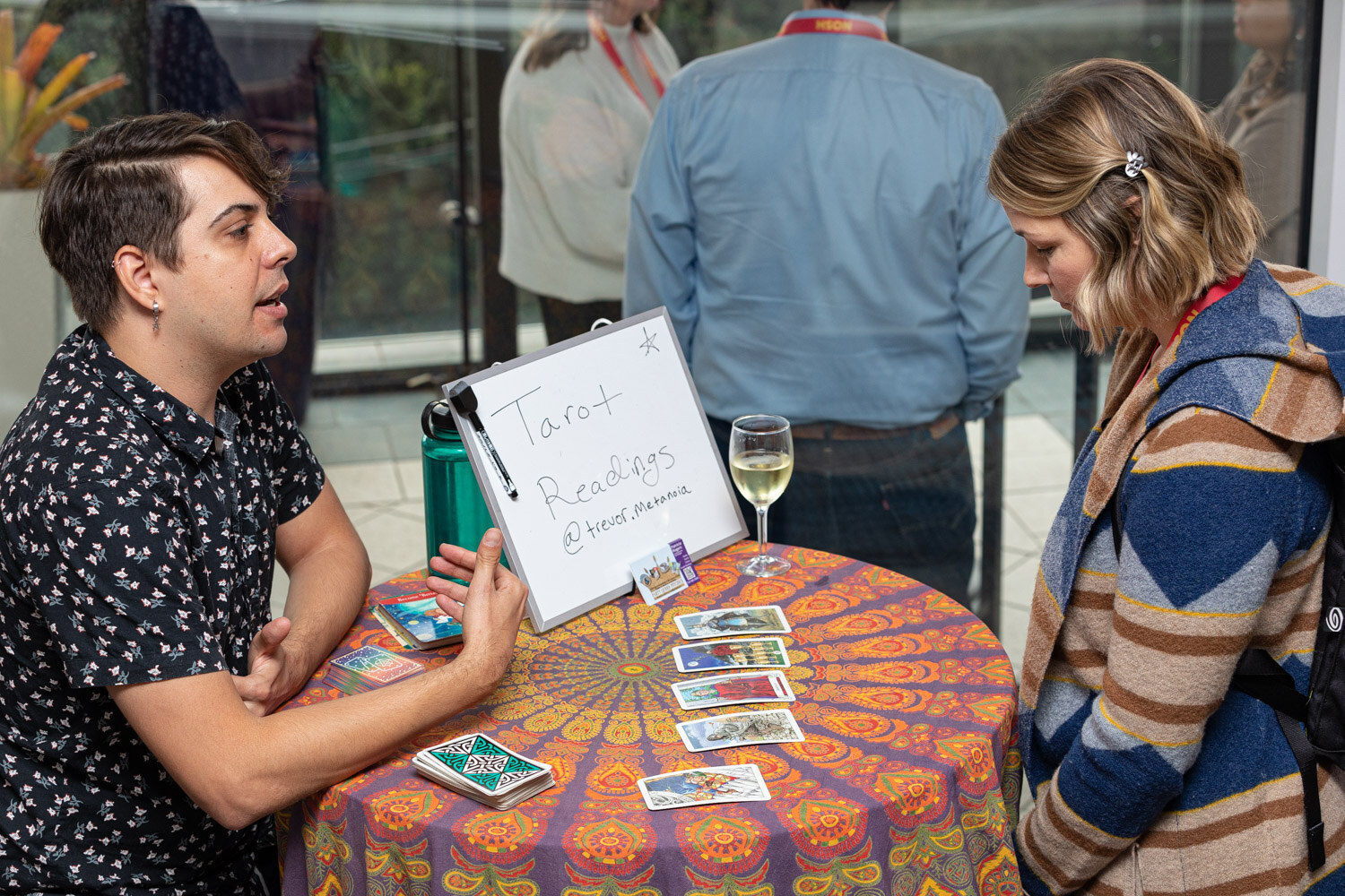 Man in a patterned shirt gesturing over tarot cards on a colorful tablecloth while a woman looks at them.