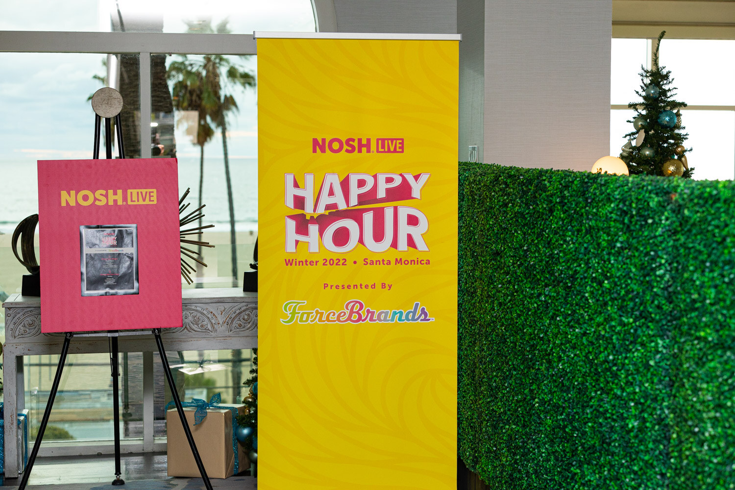 Yellow banner reading Nosh Live Happy Hour Winter 2022 Santa Monica, presented by ForceBrands, next to a pink easel.