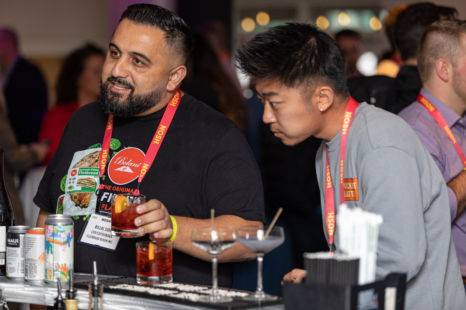 Bearded man holding a cocktail beside another man at a bar with cocktail glasses and cans.