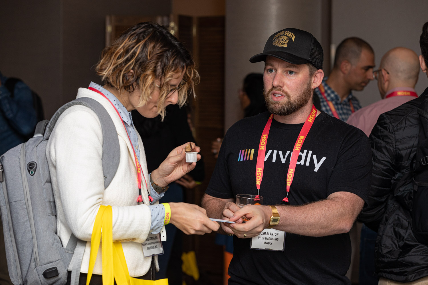 Bearded man in a black t-shirt and cap talking to a woman in a white jacket holding a small sample cup.